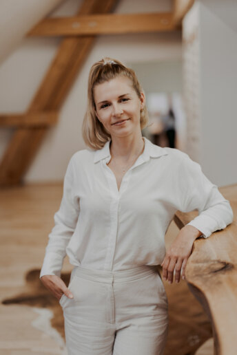 Porträt einer lächelnden Frau in weißem Outfit, stehend an einer Holztischplatte in einem hellen Raum mit sichtbaren Holzbalken.